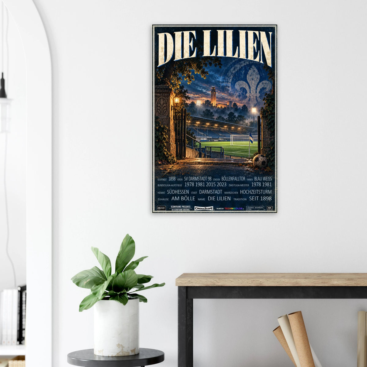 Darmstadt 98 Poster „Die Lilien“ an einer hellen Wand neben einer Pflanze und einem Tisch