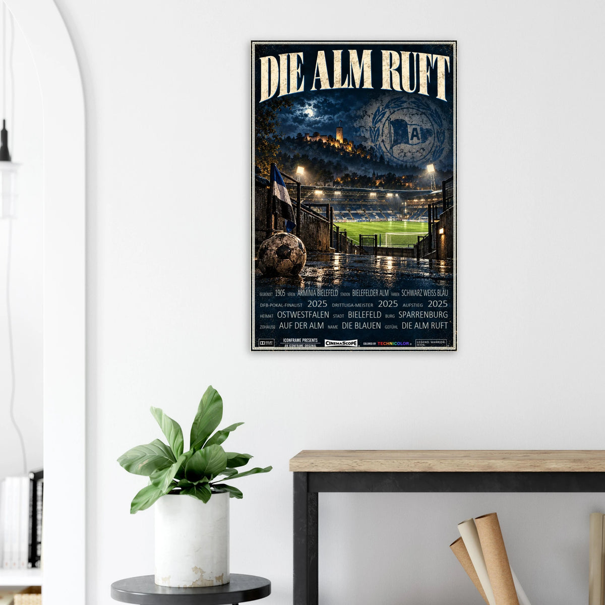 Arminia Bielefeld Poster „Die Alm ruft“ an einer hellen Wand neben einer Pflanze und einem Tisch